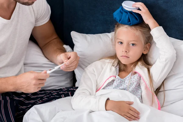 Recortado tiro de padre comprobar hijas temperatura con termómetro eléctrico mientras ella acostado con bolsa de hielo en la cama - foto de stock