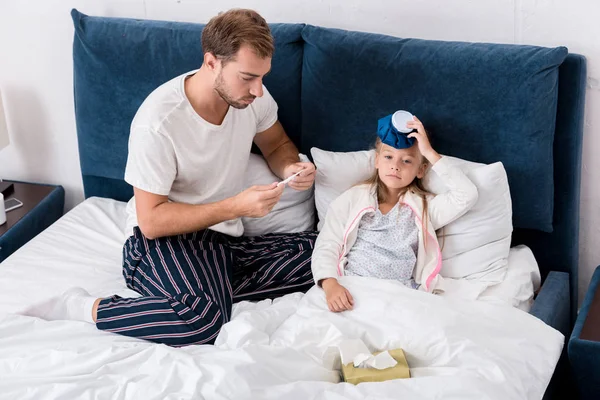 High angle view of father checking daughters temperature with electric thermometer while she lying with ice pack in bed — Stock Photo