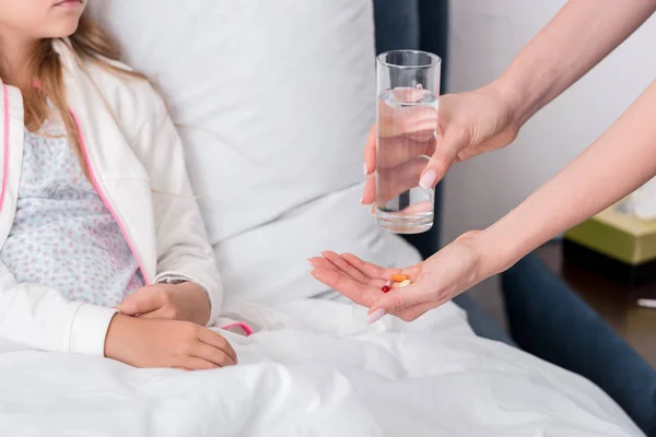 Recortado disparo de madre dando pastillas y agua a su hija enferma en la cama - foto de stock