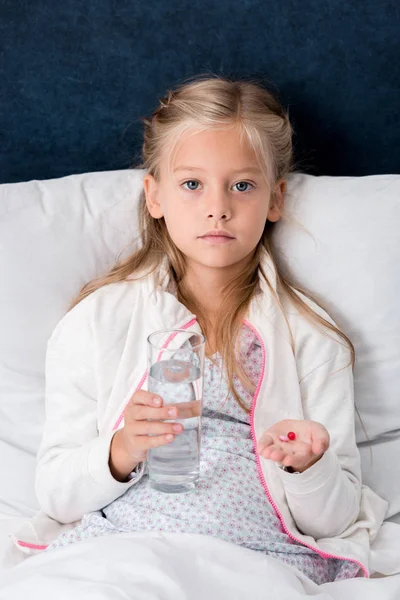 Niño enfermo con un vaso de agua y pastillas tumbado en la cama y mirando a la cámara - foto de stock