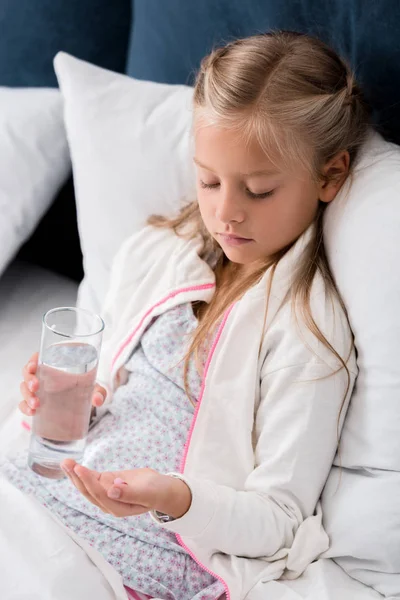 Niño enfermo con vaso de agua y pastillas acostado en la cama - foto de stock