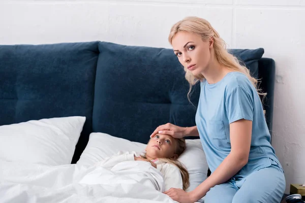 Madre joven comprobando la temperatura de sus hijas enfermas con la mano y mirando la cámara - foto de stock