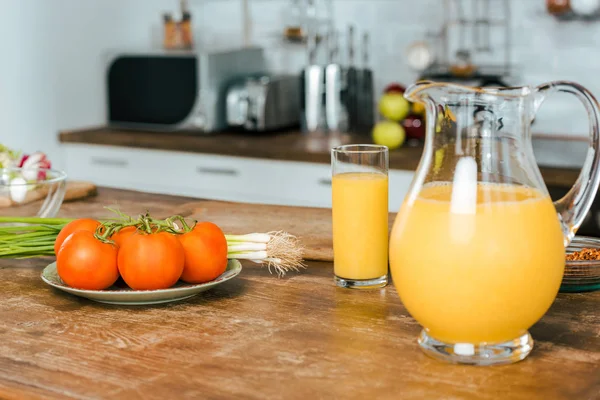 Close-up shot of raw tomatoes with leek and jug of orange juice on table at modern kitchen — Stock Photo