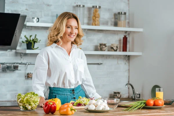 Beautiful adult woman with various fresh vegetables on kitchen table looking away — Stock Photo