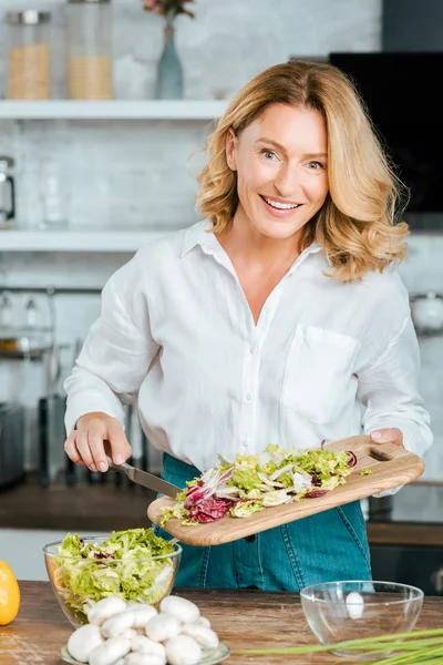 Glückliche erwachsene Frau, die in der Küche gesunden Salat zubereitet und in die Kamera schaut — Stockfoto