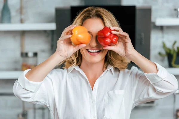 Retrato de cerca de la mujer adulta divertida cubriendo los ojos con pimientos en la cocina - foto de stock