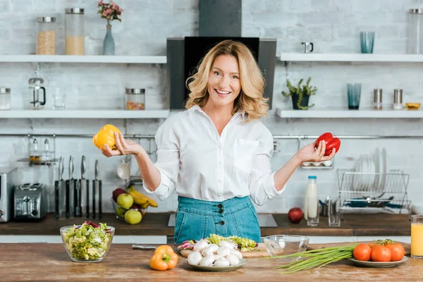 Hermosa mujer adulta con pimientos que mira a la cámara mientras hace ensalada en la cocina - foto de stock