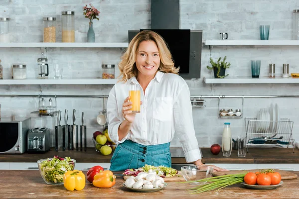 Hermosa mujer adulta bebiendo jugo de naranja mientras cocina en la cocina y mirando a la cámara - foto de stock