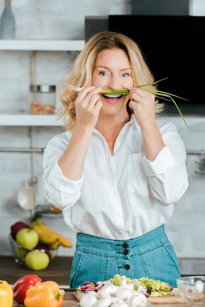 Mulher adulta engraçado anexando alho-porro como bigode na cozinha e olhando para a câmera — Fotografia de Stock