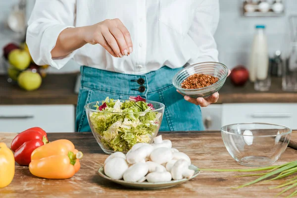 Colpo ritagliato di donna versando spezie sulla lattuga in ciotola in cucina — Foto stock