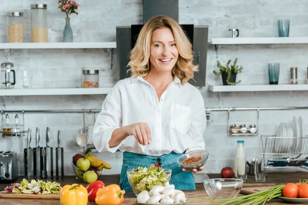 Bela mulher adulta derramando tempero na alface na tigela na cozinha e olhando para a câmera — Fotografia de Stock