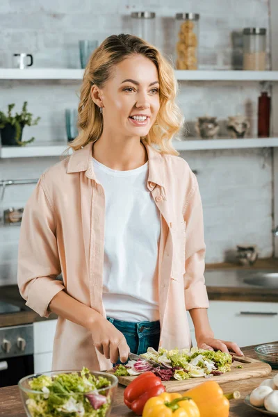 Atractiva joven corte lechuga para ensalada en la cocina y mirando hacia otro lado - foto de stock