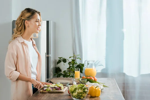 Vista lateral de atraente jovem mulher cortando alface para salada na cozinha — Fotografia de Stock
