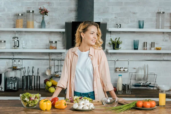 Attractive young woman with various ripe vegetables on table looking away at kitchen — Stock Photo