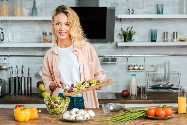 Glückliche junge Frau mit Schneidebrett vom geschnittenen Salat in der Küche — Stockfoto