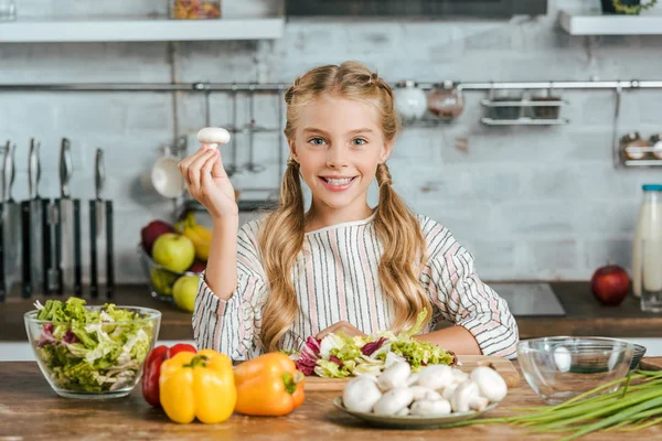 Schönes kleines Kind mit Pilzen und verschiedenem rohen Gemüse, das in die Kamera schaut, während es in der Küche Salat zubereitet — Stockfoto