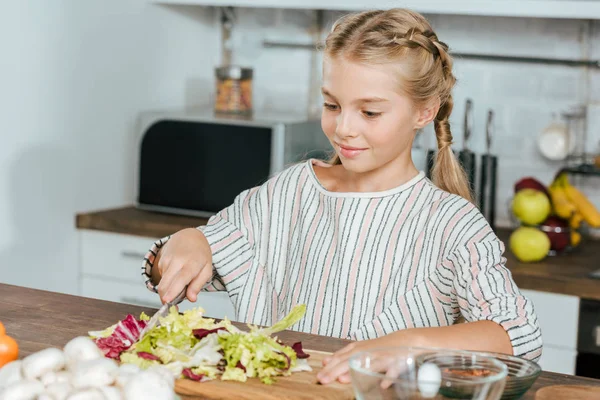 Beautiful little child cutting lettuce for salad at kitchen — Stock Photo