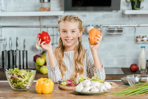 Lächelndes kleines Kind mit Paprika, das in die Kamera schaut, während es in der Küche Salat zubereitet — Stockfoto