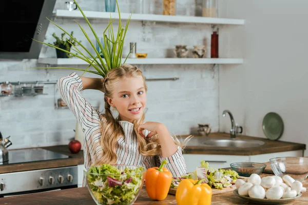 Sorrindo criança segurando monte de alho-porro atrás da cabeça ao fazer salada na cozinha e olhando para a câmera — Fotografia de Stock