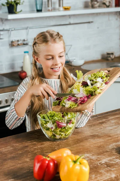 Lächelndes kleines Kind gießt geschnittenen Salat in Schüssel für Salat in der Küche — Stockfoto