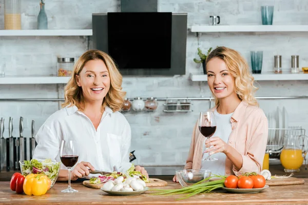 Mature mère et fille adulte avec des verres de vin rouge cuisine ensemble et regardant la caméra à la cuisine — Photo de stock