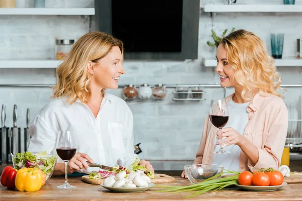 Madre madura e hija adulta con vasos de vino tinto cocinando juntos y charlando en la cocina - foto de stock