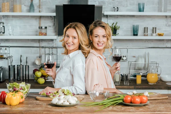 Madre madura e hija adulta con copas de vino tinto de pie espalda con espalda en la cocina y mirando a la cámara - foto de stock