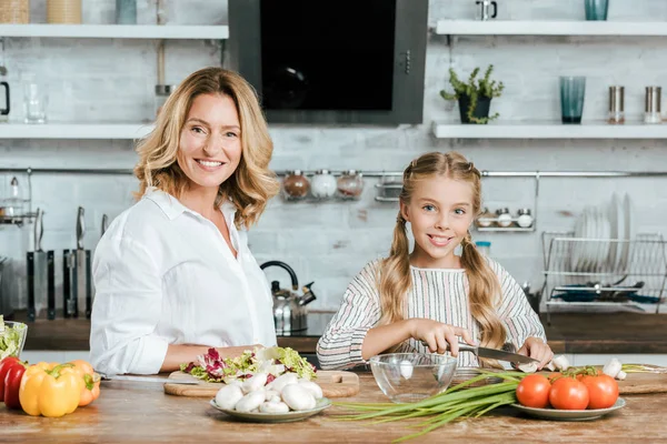 Sourire adulte mère et petite fille cuisiner ensemble et regarder la caméra — Photo de stock