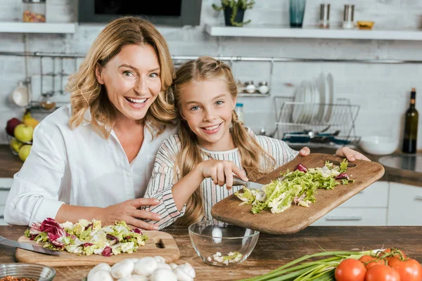 Счастливая взрослая мать и маленькая дочь делают салат вместе дома и смотрят в камеру — стоковое фото