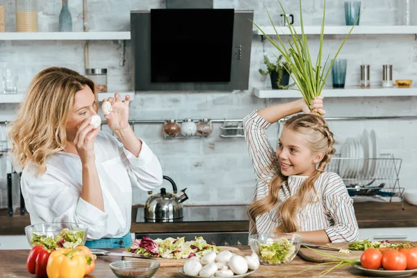 Улыбающиеся мать и дочь играют с едой во время приготовления пищи на глядя друг на друга дома — стоковое фото