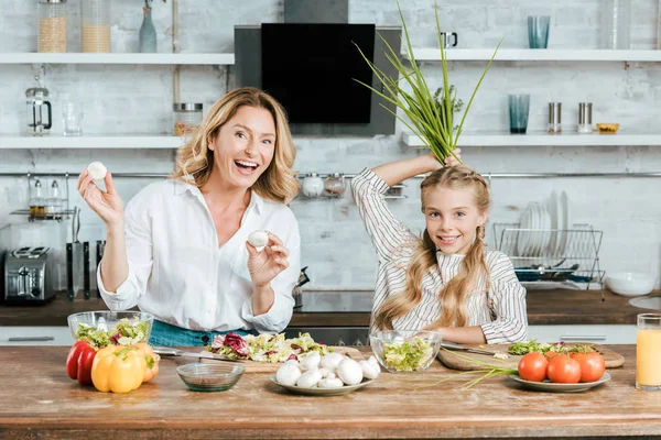 Heureux mère et fille jouer avec la nourriture tout en cuisinant à regarder la caméra à la maison — Photo de stock