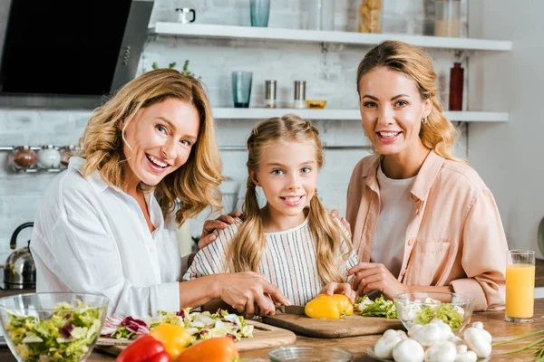 Happy little child cutting vegetables for salad with mother and grandmother at home and looking at camera — Stock Photo