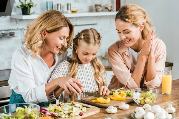 Adorable little child cutting vegetables for salad with mother and grandmother at home — Stock Photo