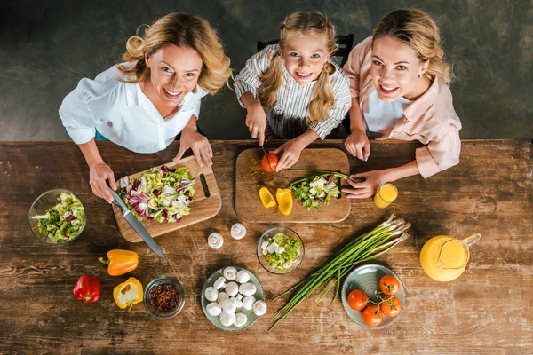 Overhead view of child cutting vegetables for salad with mother and grandmother and looking at camera — Stock Photo