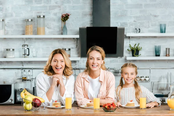 Laughing three generations of women with pancakes and orange juice for breakfast at home looking at camera — Stock Photo