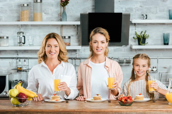 Belas três gerações de mulheres com panquecas e suco de laranja para o café da manhã em casa olhando para a câmera — Fotografia de Stock