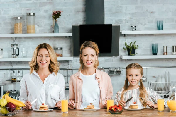 Beautiful three generations of women with pancakes for breakfast at home looking at camera — Stock Photo