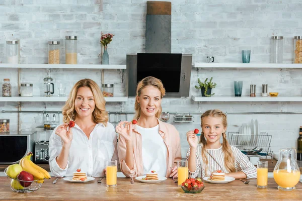 Smiling three generations of women with pancakes and strawberries for breakfast at home looking at camera — Stock Photo