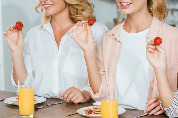 Foto recortada de mujeres sonrientes sosteniendo fresas mientras desayunan con panqueques y jugo de naranja - foto de stock