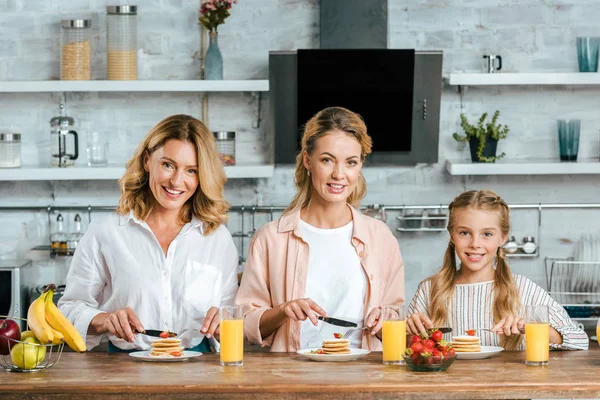 Beautiful child with mother and grandmother eating pancakes for breakfast at home looking at camera — Stock Photo