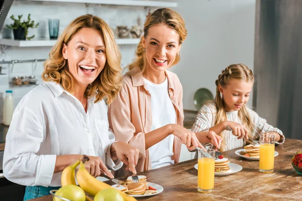 Feliz tres generaciones de mujeres tomando panqueques para el desayuno en casa mirando a la cámara - foto de stock