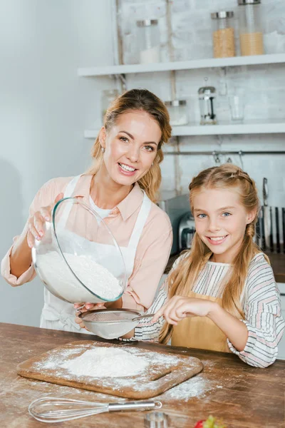 Sorrindo mãe e filha derramando farinha através da peneira para massa na cozinha e olhando para a câmera — Fotografia de Stock