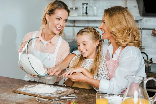 Happy child with mother and grandmother pouring flour through sieve during dough preparation at home — Stock Photo