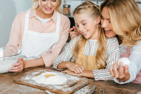 Close-up shot of little child with mother and grandmother making dough with flour and egg together at home — Stock Photo