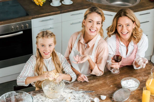 Высокий угол обзора улыбающегося ребенка с мамой и бабушкой, которые вместе делают тесто дома и смотрят в камеру — стоковое фото