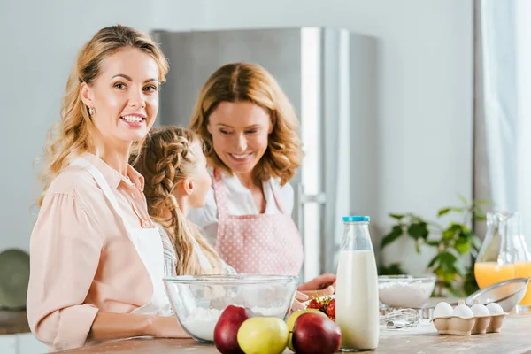 Красивая молодая женщина готовит дома с мамой и дочерью и смотрит в камеру — стоковое фото