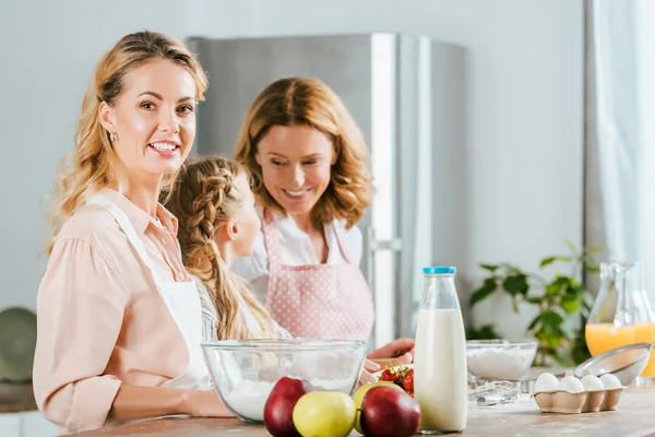 Bella giovane donna che cucina con la madre e la figlia a casa e guardando la fotocamera — Foto stock