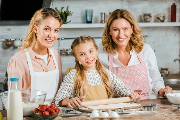 Красивые улыбающиеся три поколения женщин, которые вместе делают тесто дома и смотрят в камеру — стоковое фото
