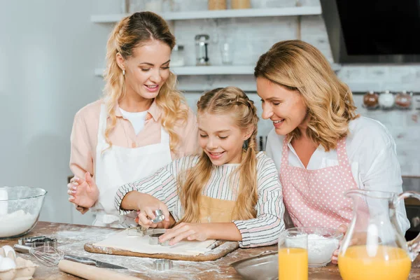Улыбающийся маленький ребенок режет тесто для печенья с мамой и бабушкой дома — стоковое фото