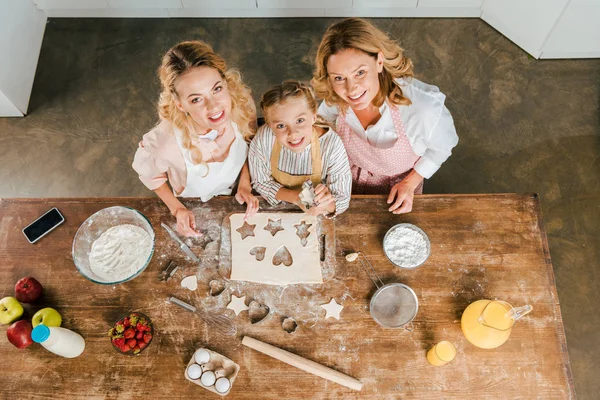 Vista superior de criança sorridente com mãe e avó preparando biscoitos de Natal em casa e olhando para a câmera — Fotografia de Stock